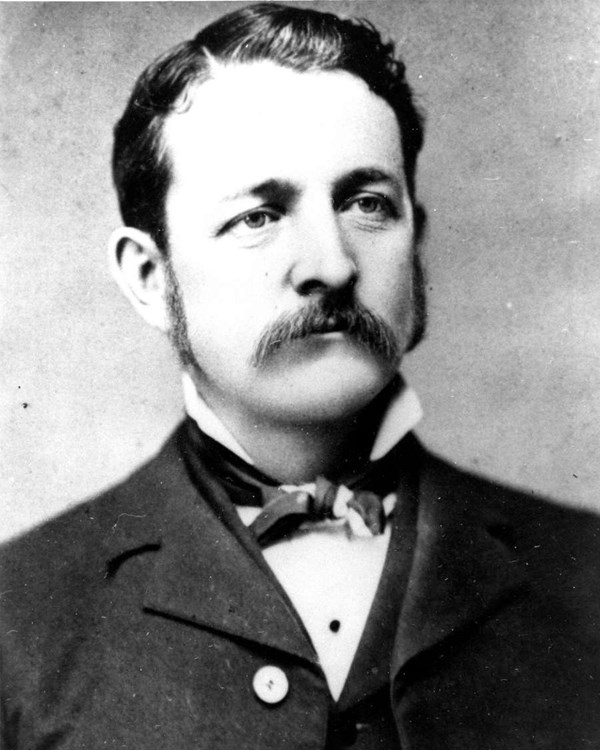 An 1883 portrait photo depicts a man with a mustache and sideburns in a suit with a bow tie.