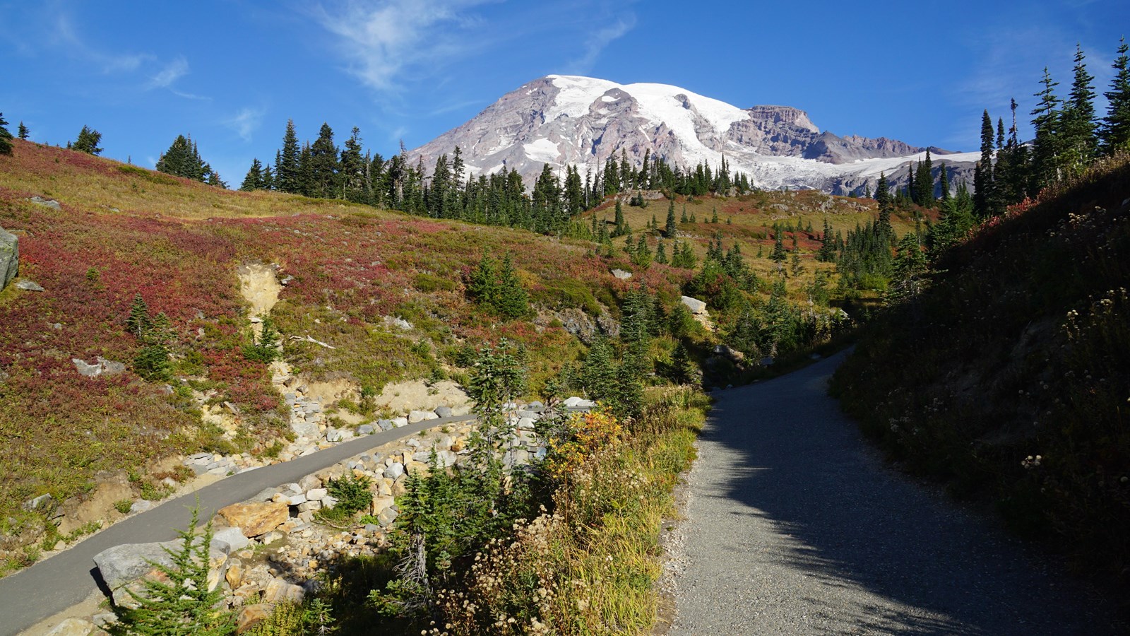 A glaciated mountain rises above autumn meadows and a paved trail intersection. 