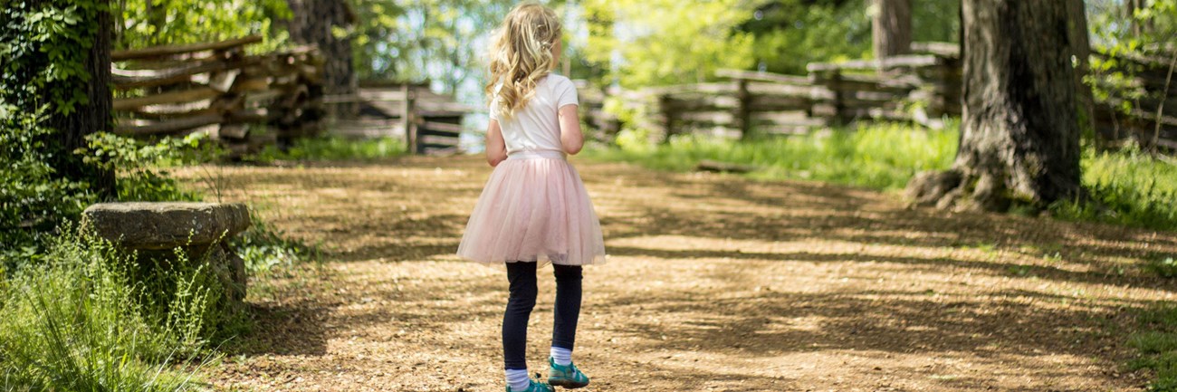 young girl walks on trail lined by a split-rail fence.