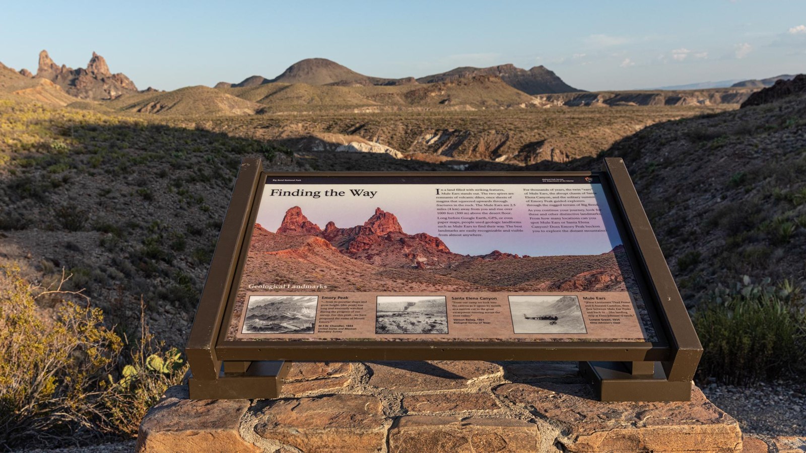 A sign sits on a rock mount with a view looking out towards the twin spires of Mule Ears.