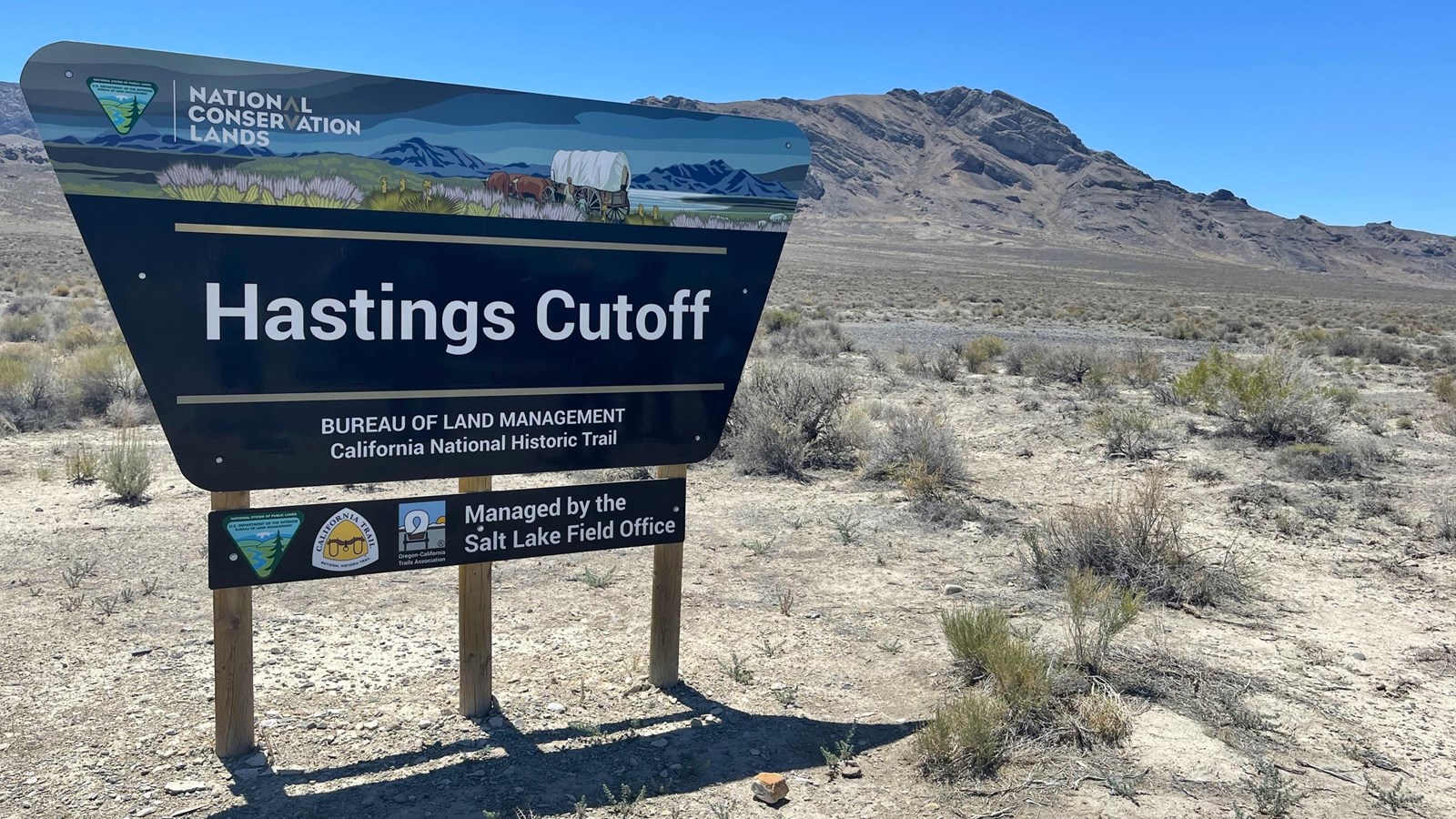 BLM sign marks Hastings Cutoff amid desert shrubs and distant rocky mountains under blue sky.