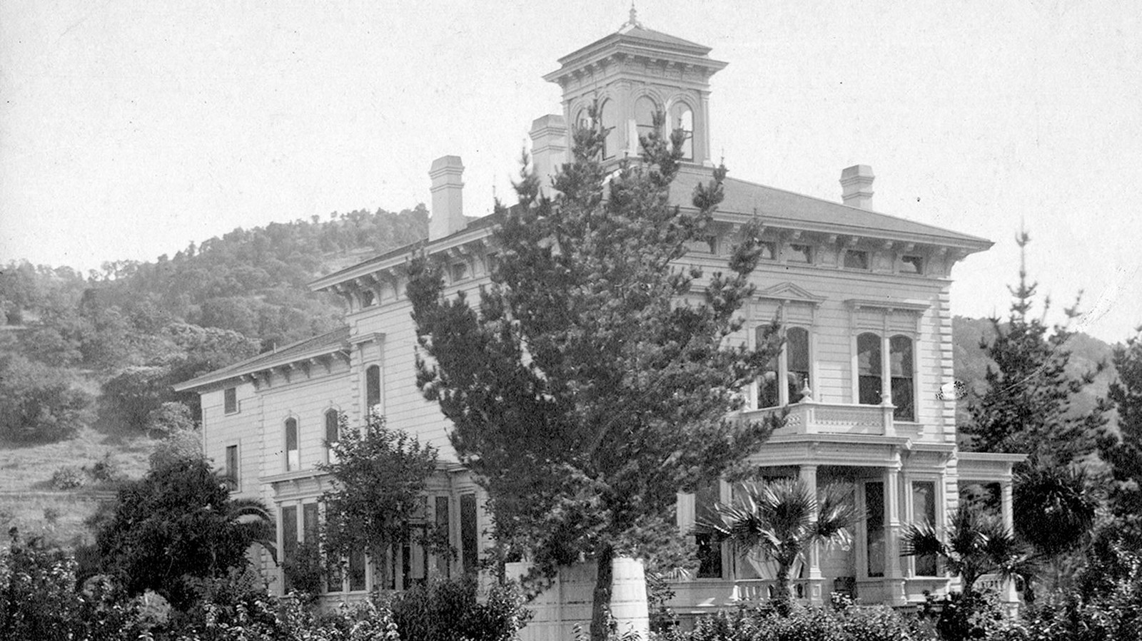 An historic image of a two-story victorian home, surrounded by trees and bushes. 