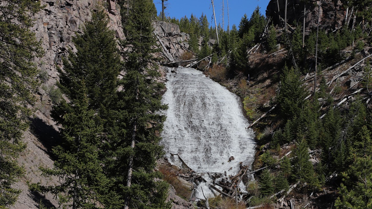 Water cascades down a slope surrounded by rocky cliffs and sparse trees.