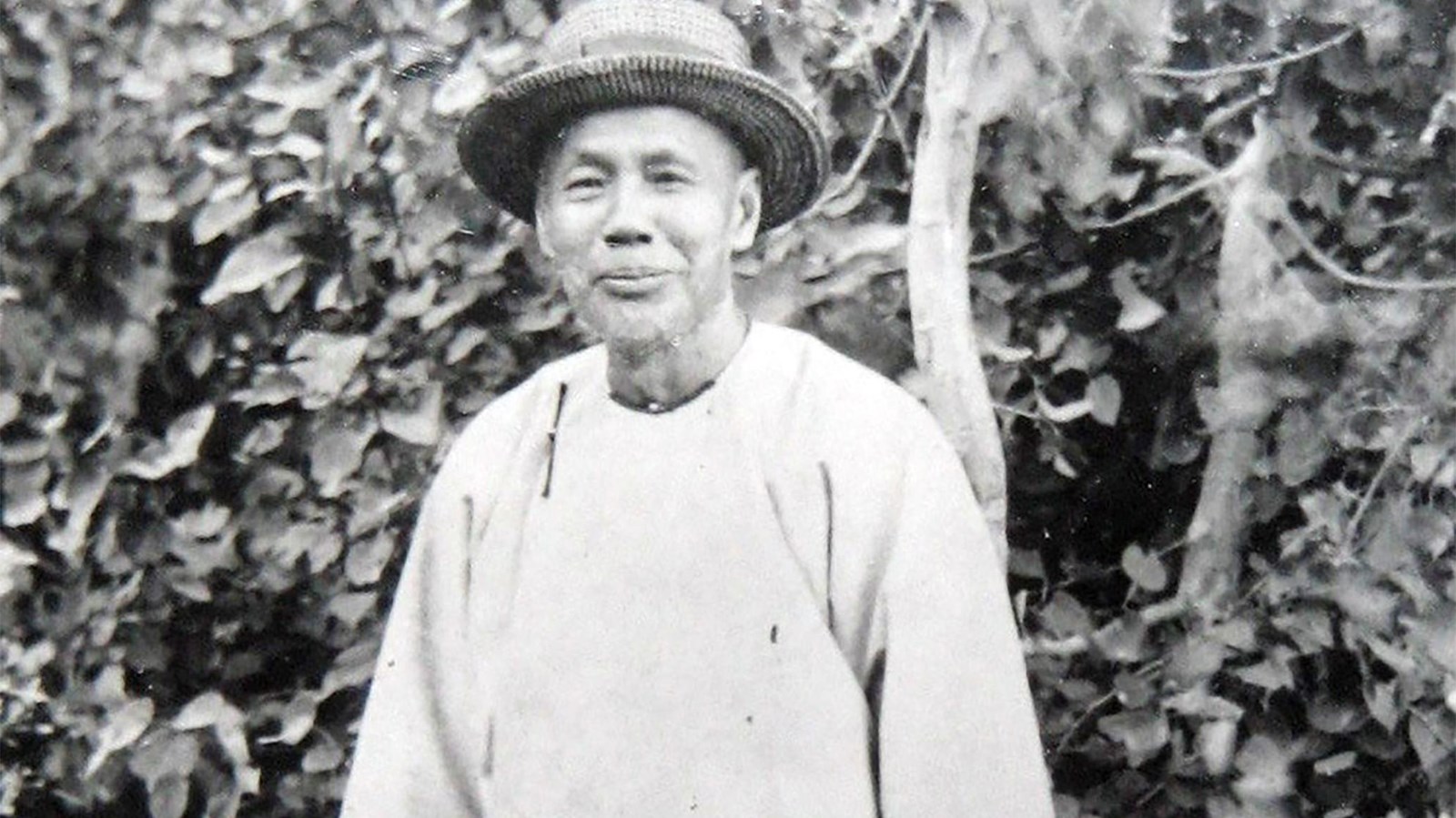 An older Chinese male is holding a basket and standing in front of some trees. He is smiling. 