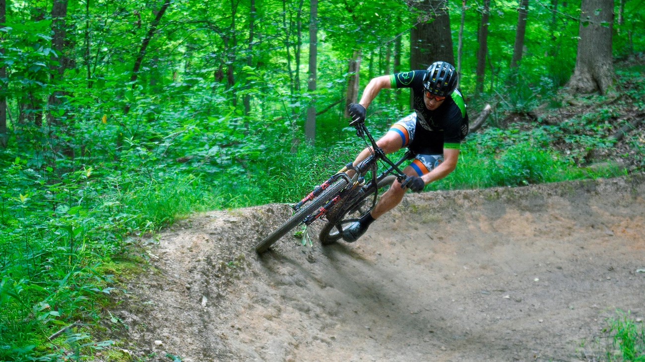 A mountain biker wearing a helmet banks around a curve at a 45-degree angle, on a wooded trail.