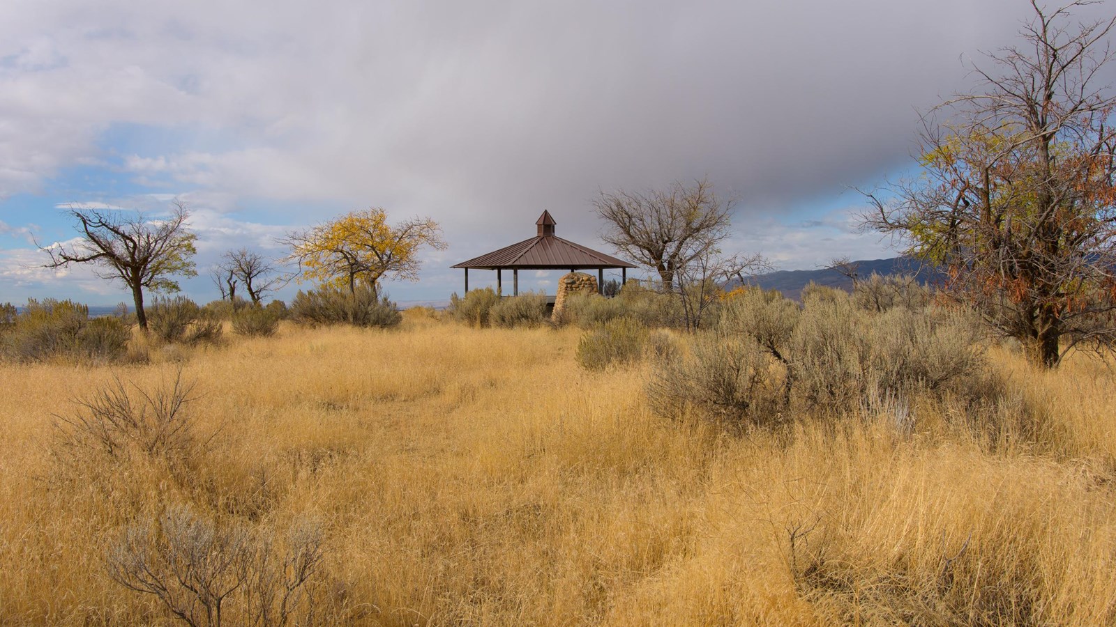 A picnic pavilion sits on top of a rounded hill covered with warm-colored grass.