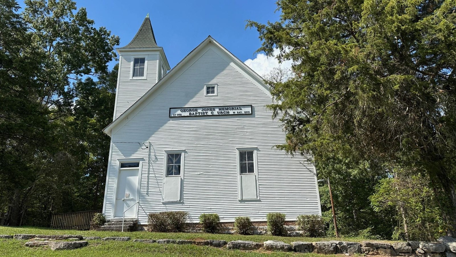 A large white church sits on a grassy hill surrounded by woods.