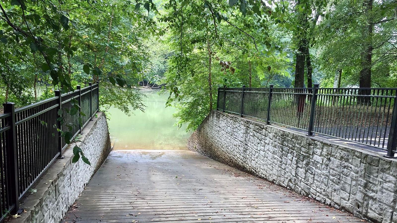 Looking down paved boat ramp with fence topped walls on either side. Wooded area behind walls.