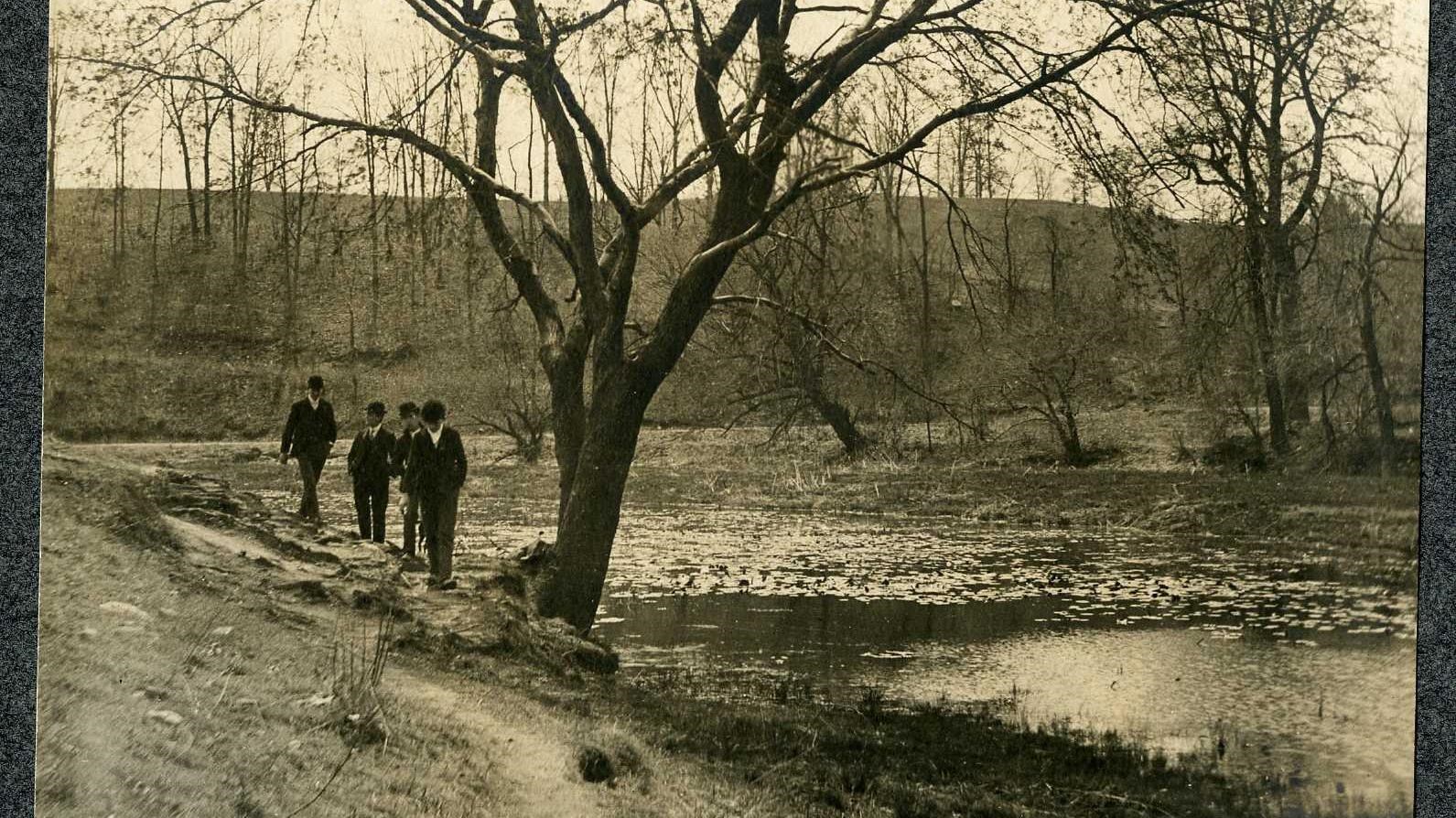 Black and white of four men on side of water walking on dirt path with trees around