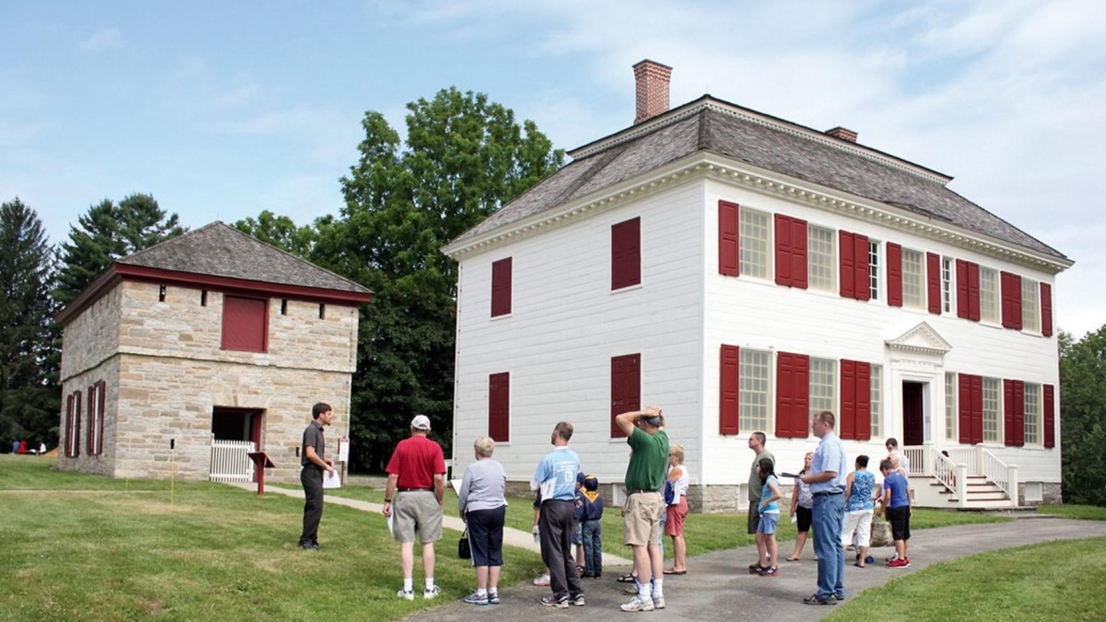 A group of people stand behind a large, whitewashed house with red shutters.