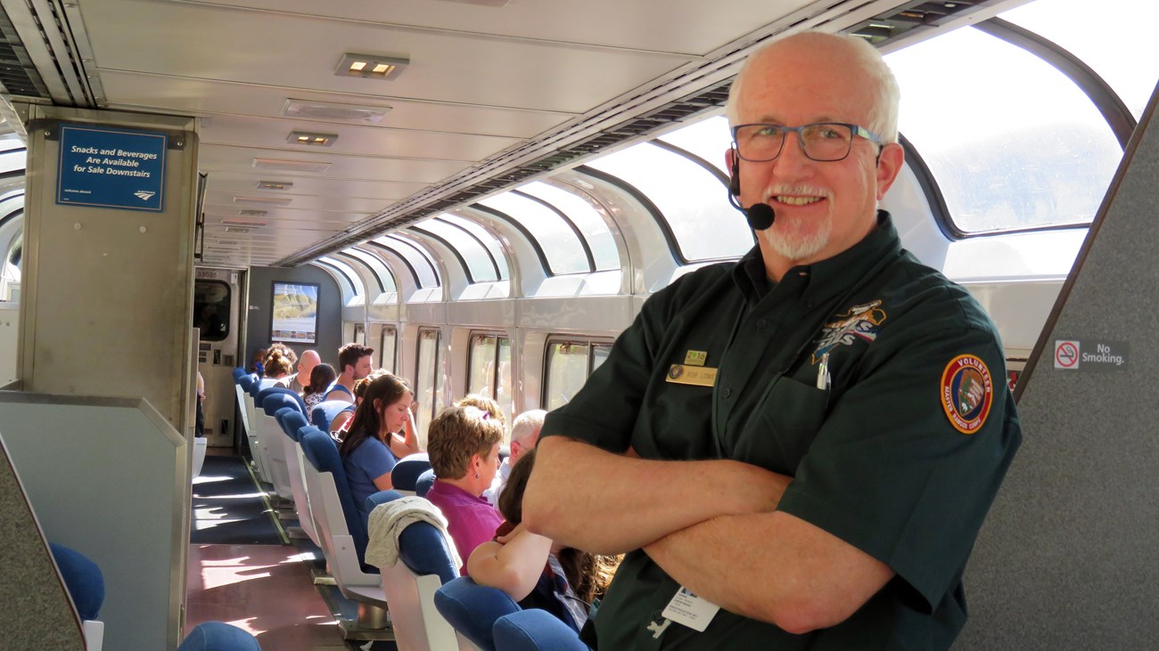 Man in green shirt poses in foreground of an Amtrak sightseer train car.