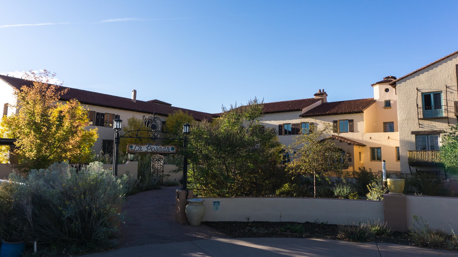 A cream colored two story adobe style building with a brown roof, surrounded by trees.