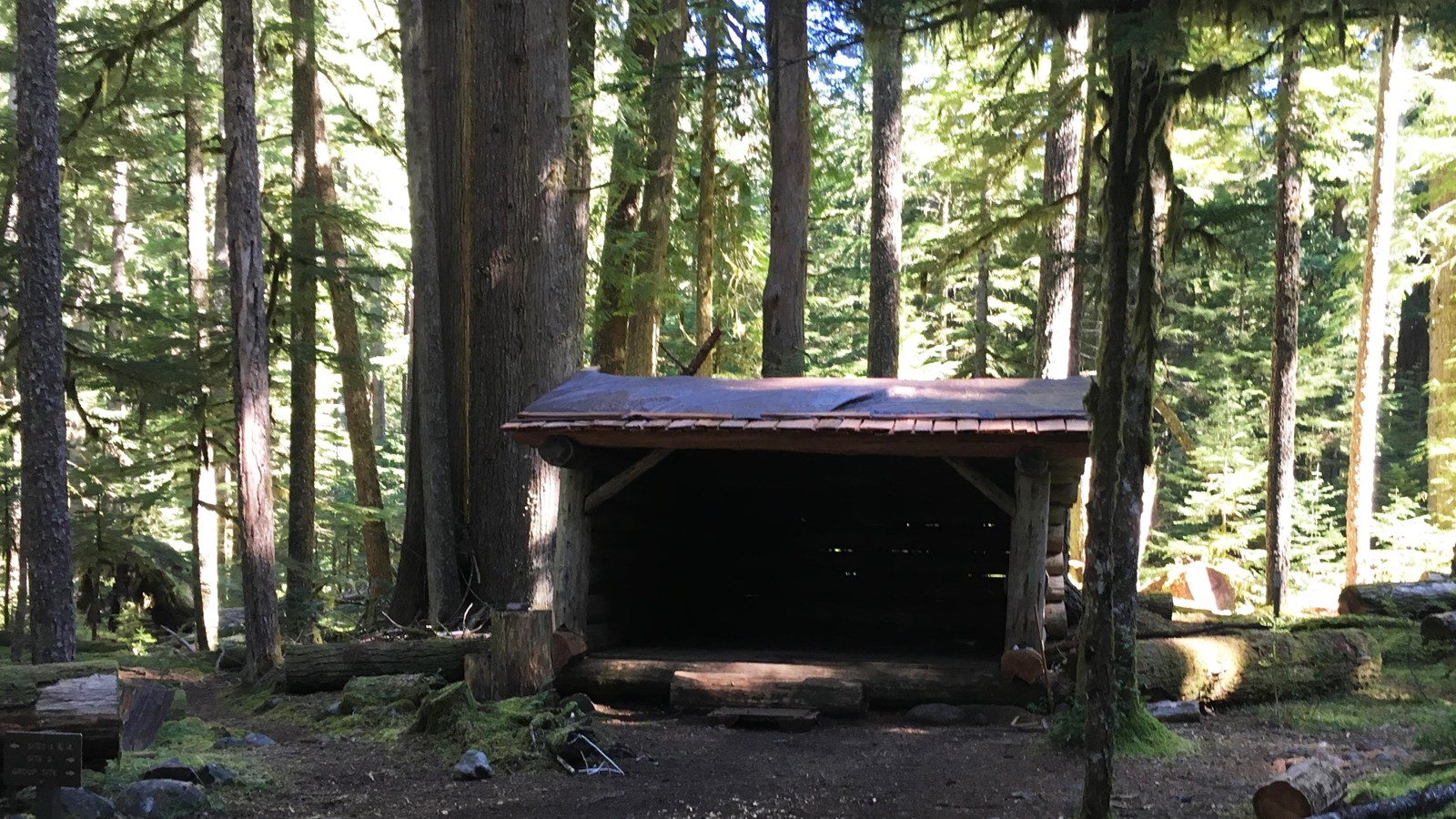 A small log trail shelter with three sides and open front side, covered by a shingle roof.