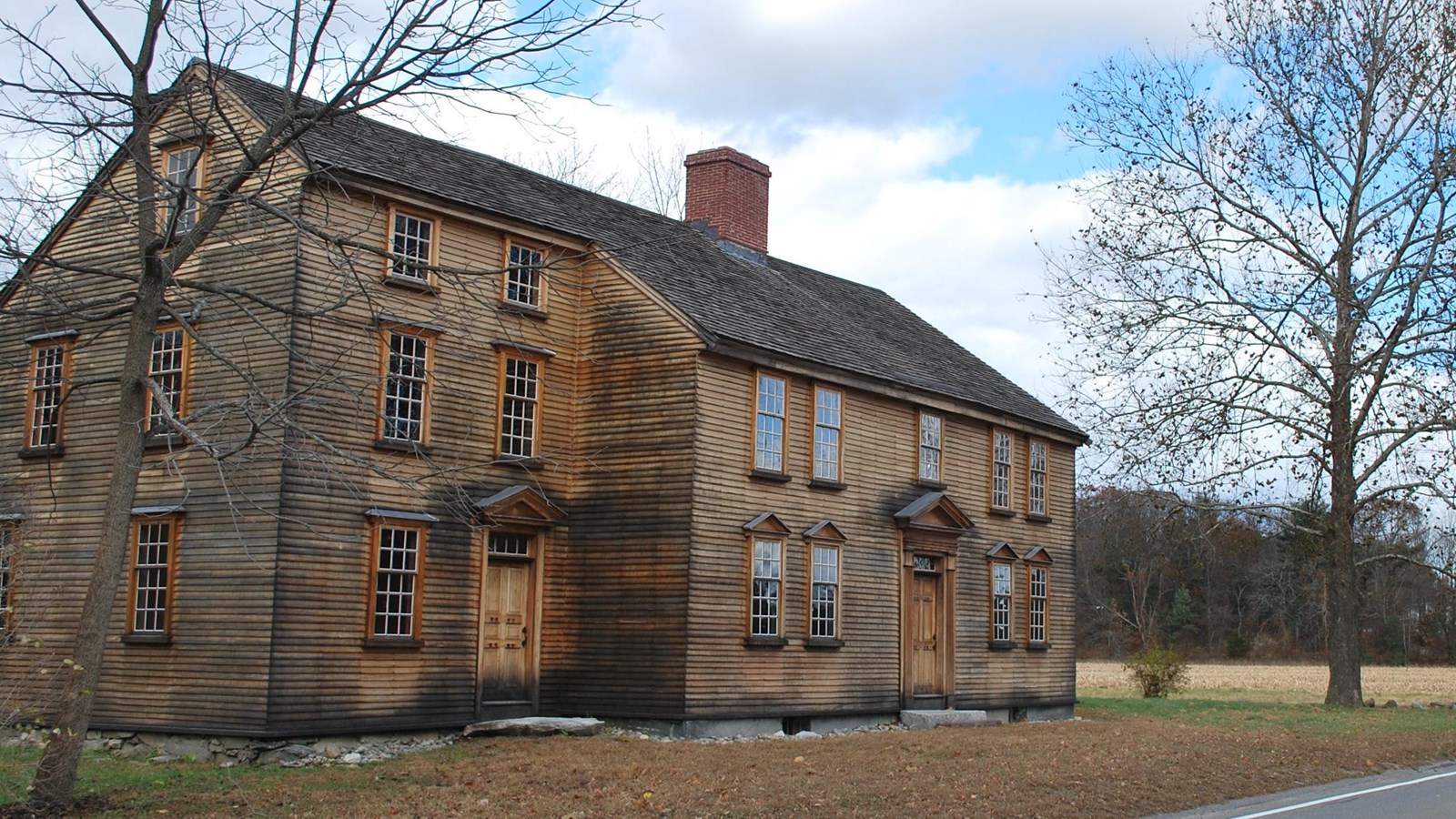 Two story wooden colonial house, unpainted wooden siding and a central chimney. 