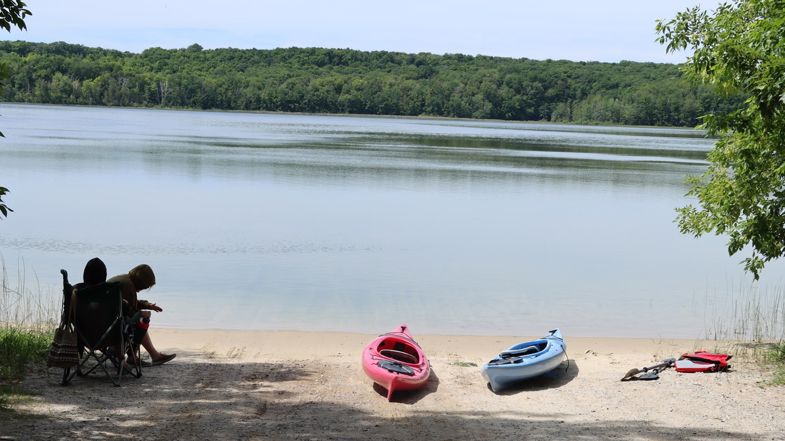 Two people rest in the shade near the lake, two kayaks with paddles and life jackets dry in the sun.