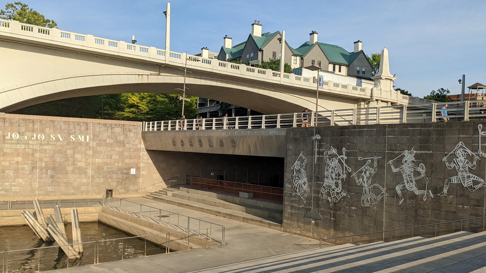 Concrete walkway along waterway. Artistic Cherokee figures painted on dark block wall. 