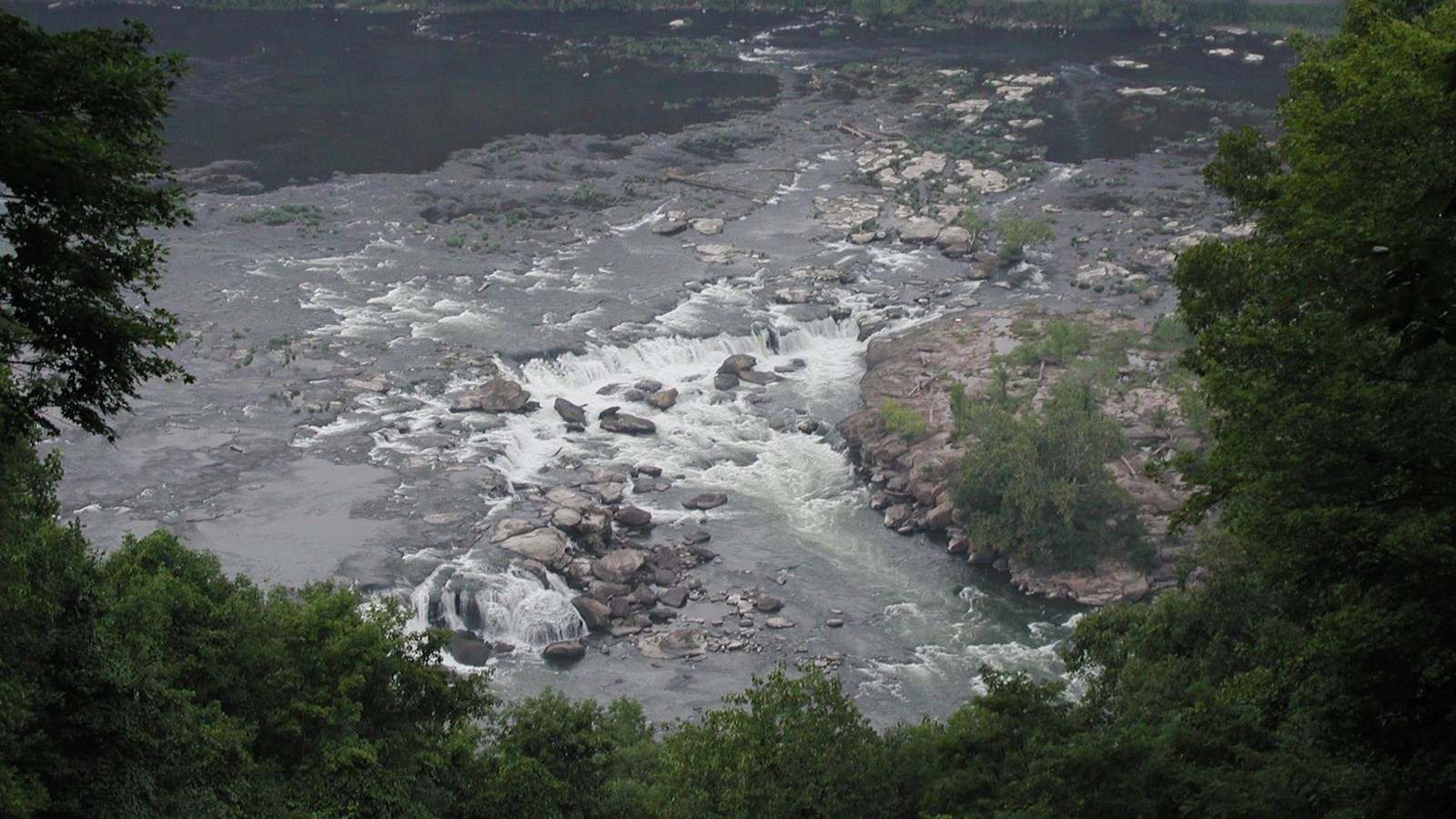 Looking down onto the river and a waterfall through bright green trees.