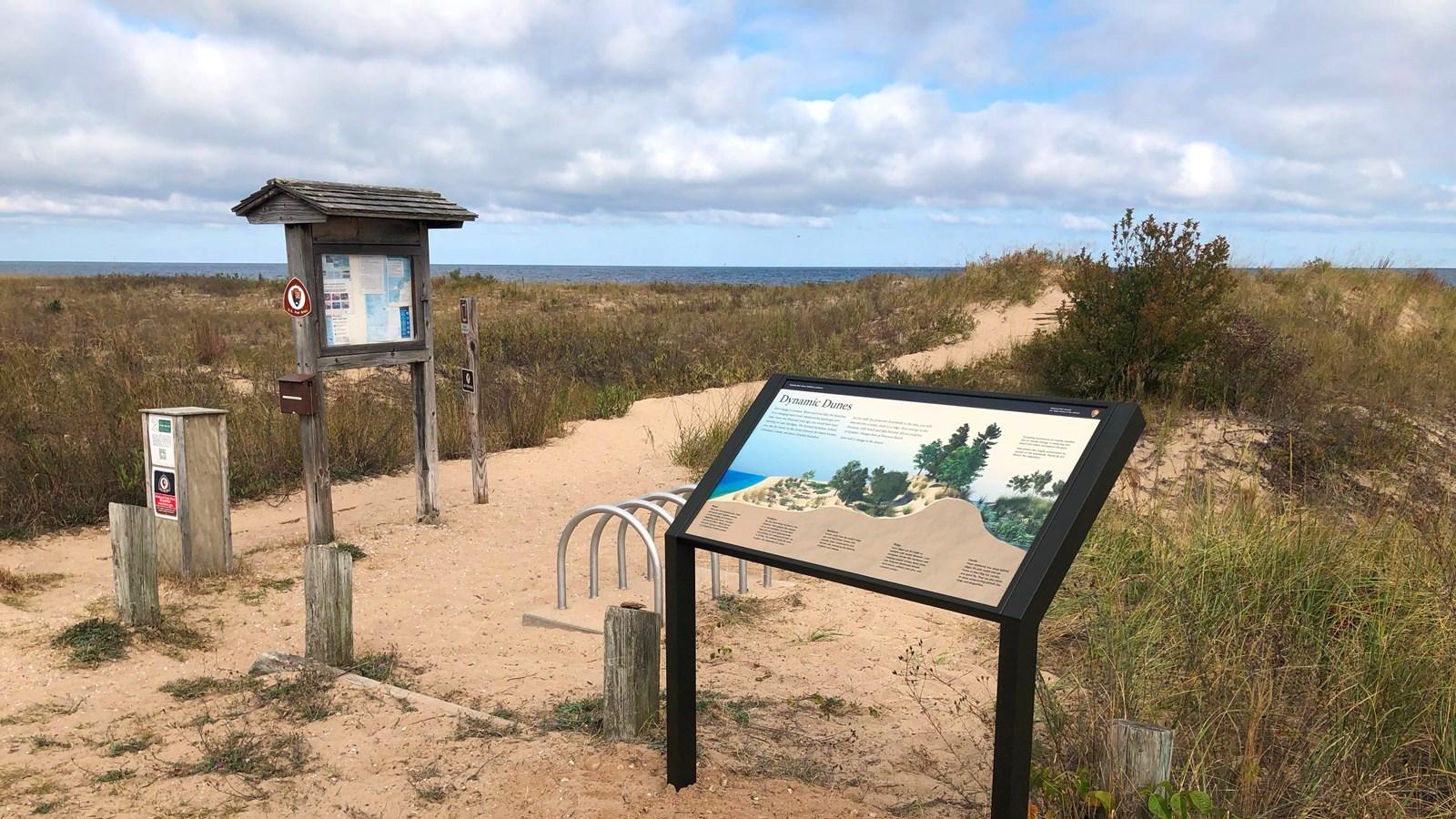 An informational sign stands in front of a bike rack at the start of a sandy path lined with grasses