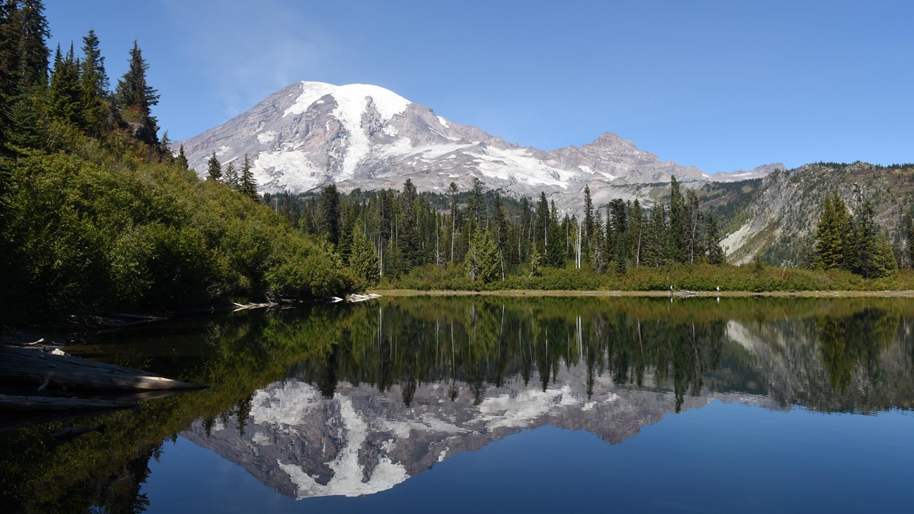 Glaciated mountain with mirrored reflection in lake with blue sky and trees