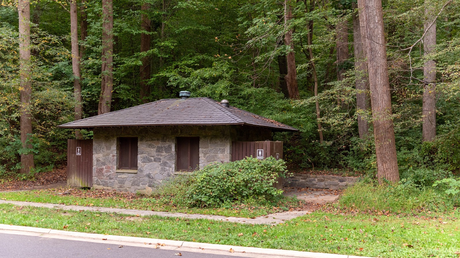 A stone restroom in a wooded area next to a road.  