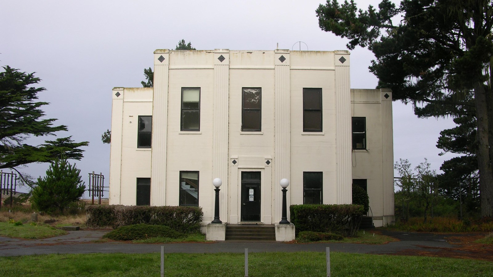 A white two-story art deco building flanked by trees beyond a green roundabout.