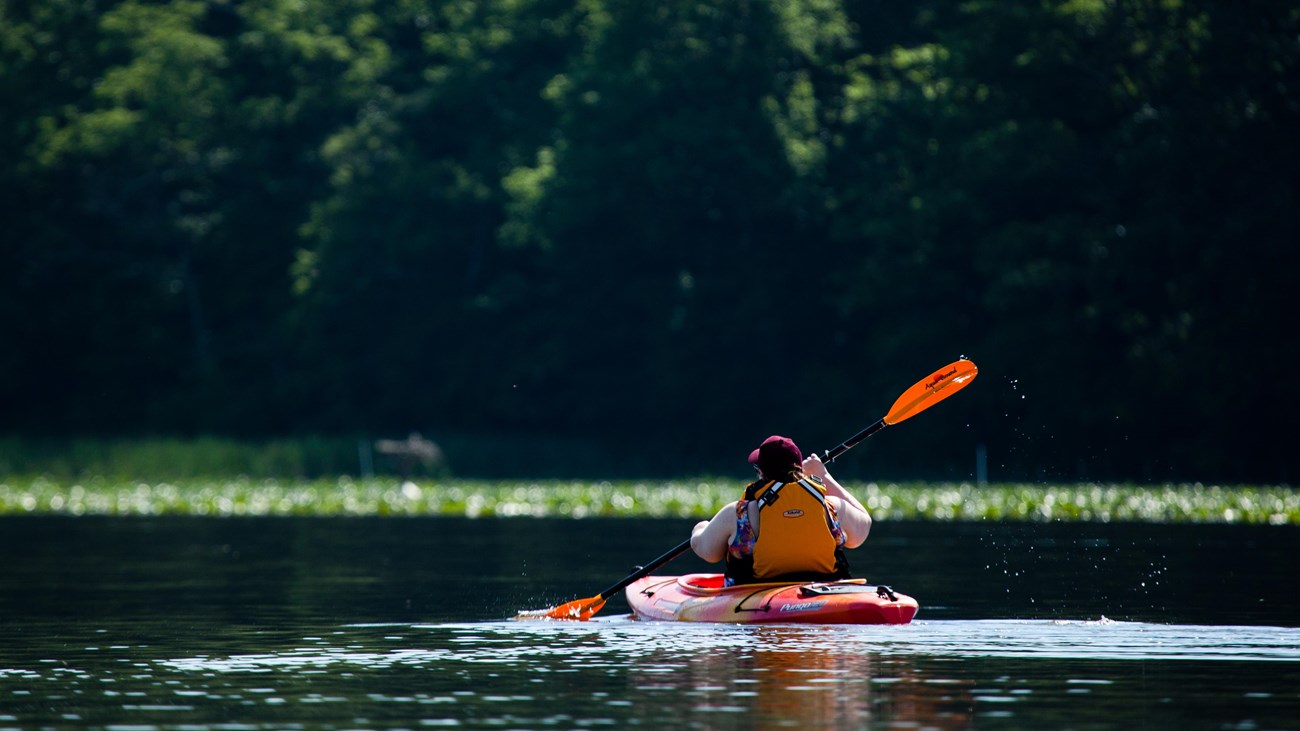 A person paddles an orange kayak on a river with trees in the background.