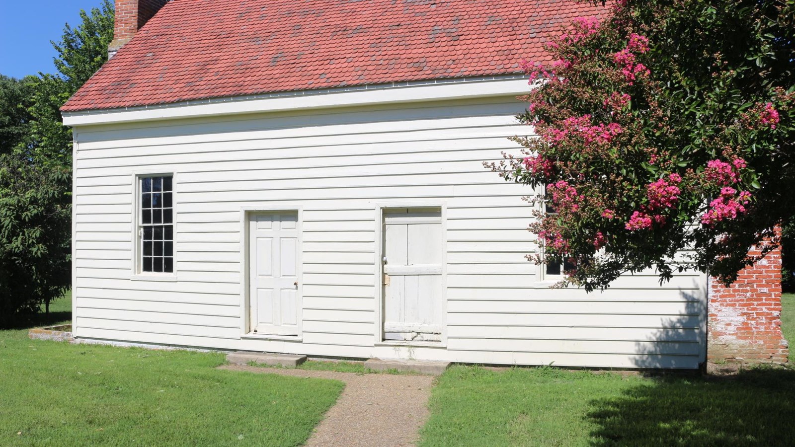 White two-story building with red roof and two red chimneys, tree in foreground of image.