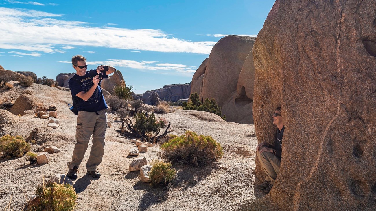 A man on a trail holds a camera and smiles towards a person kneeling in front of a rock