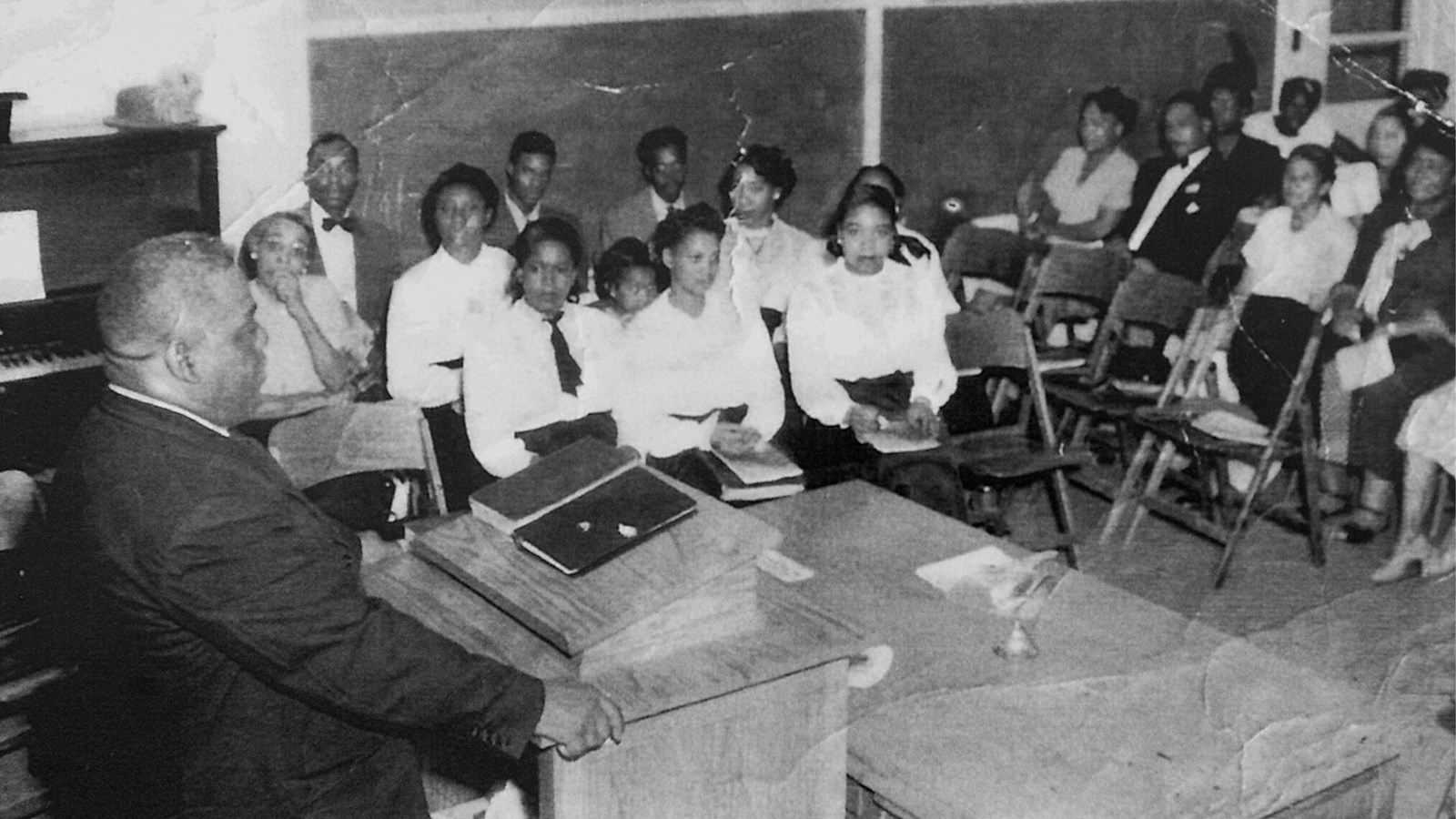 A Black minister stands at a podium while over a dozen people seated in chairs look toward him.