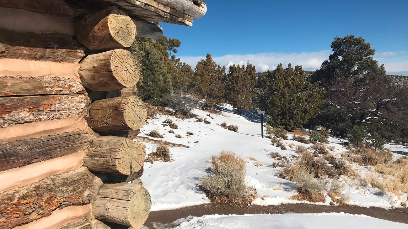 The edge of a log cabin with large beams sits to one side, with a trail stretching forward in snow