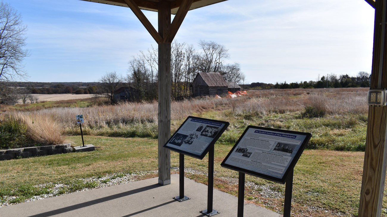 Two information signs stand at the edge of a concrete floor, which overlooks fields and old cabins.