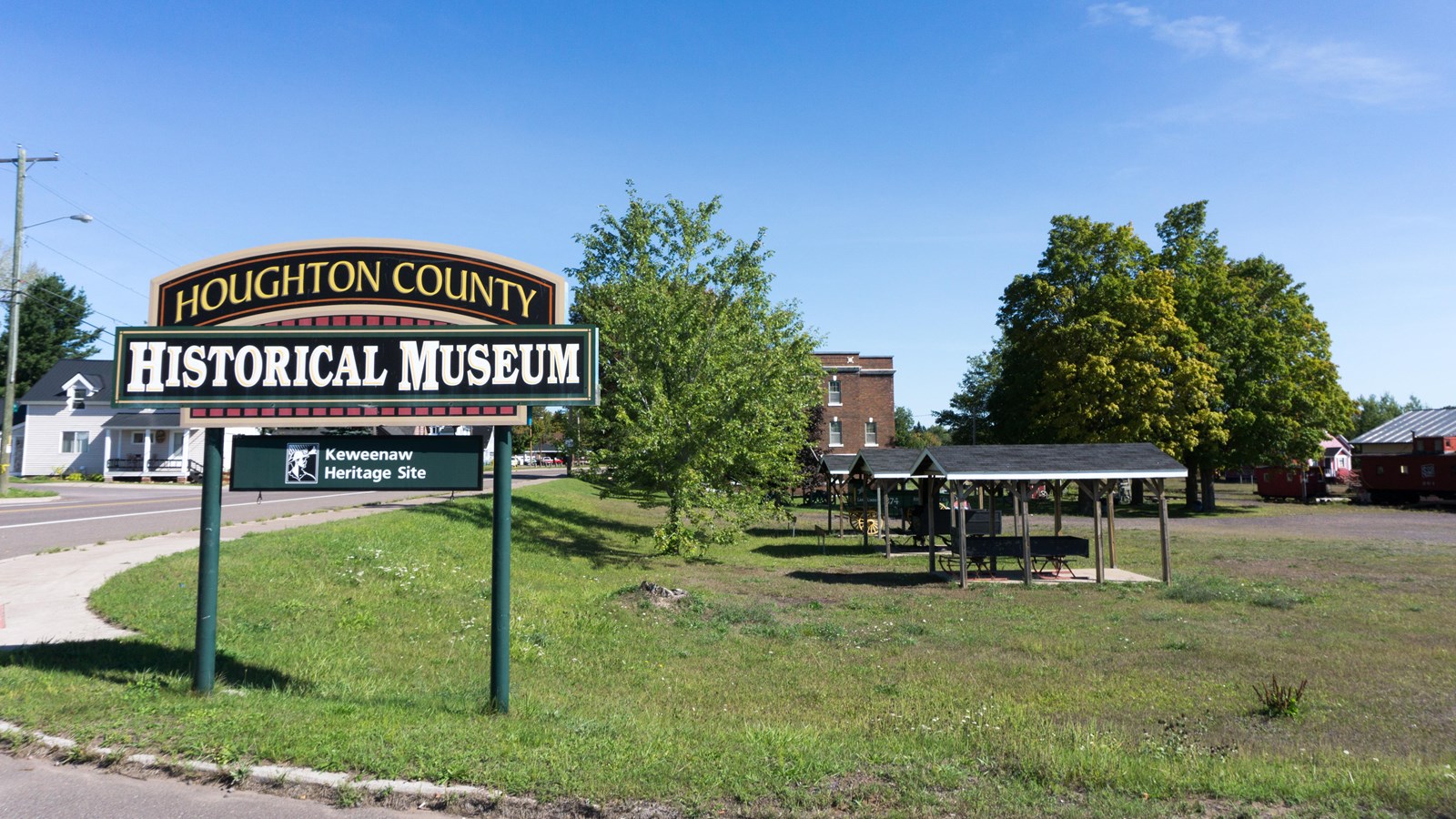 Summer scene of sign:Houghton County Historical Museum with covered antique slays in the background.