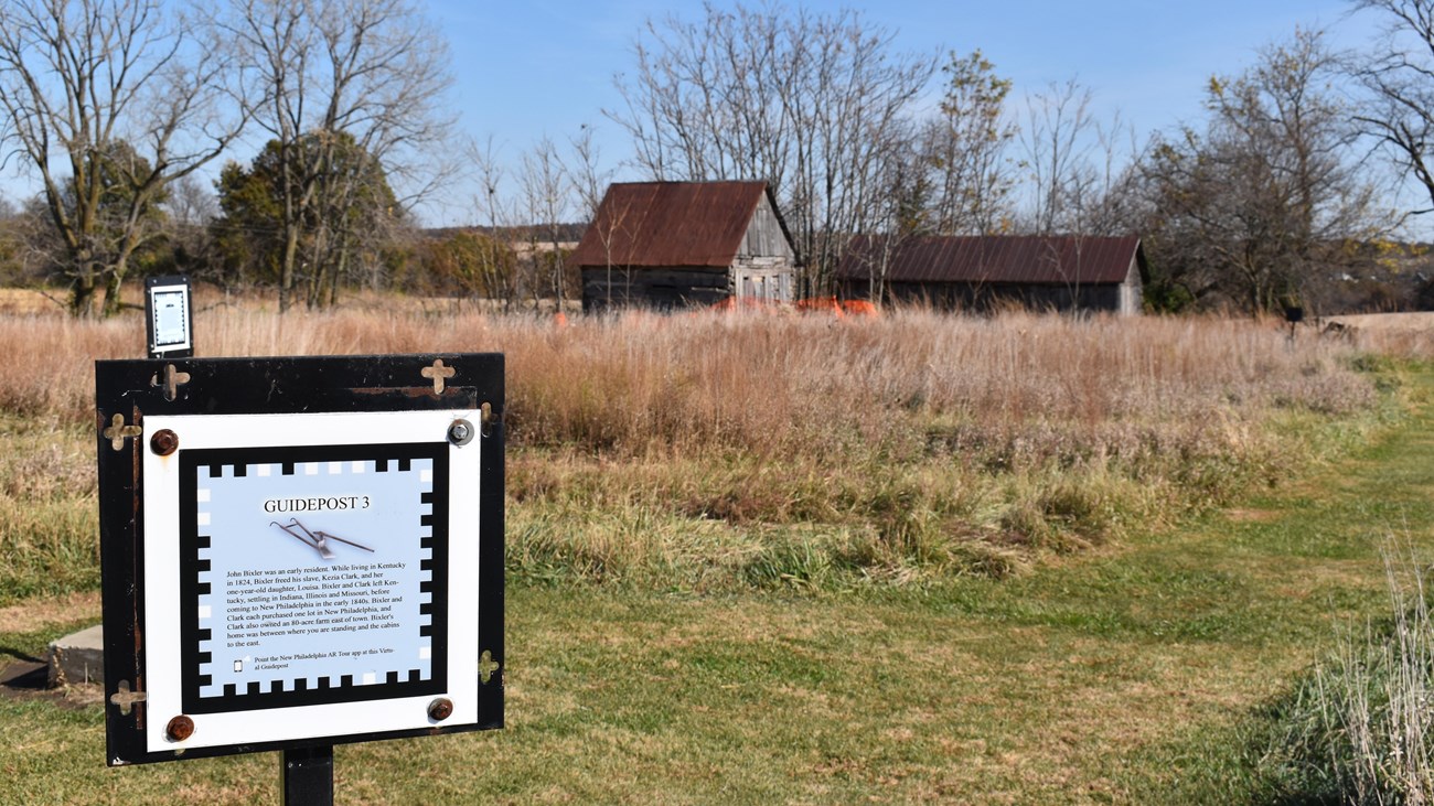 An information sign with QR code stands in front of historic wood cabins in the distance.