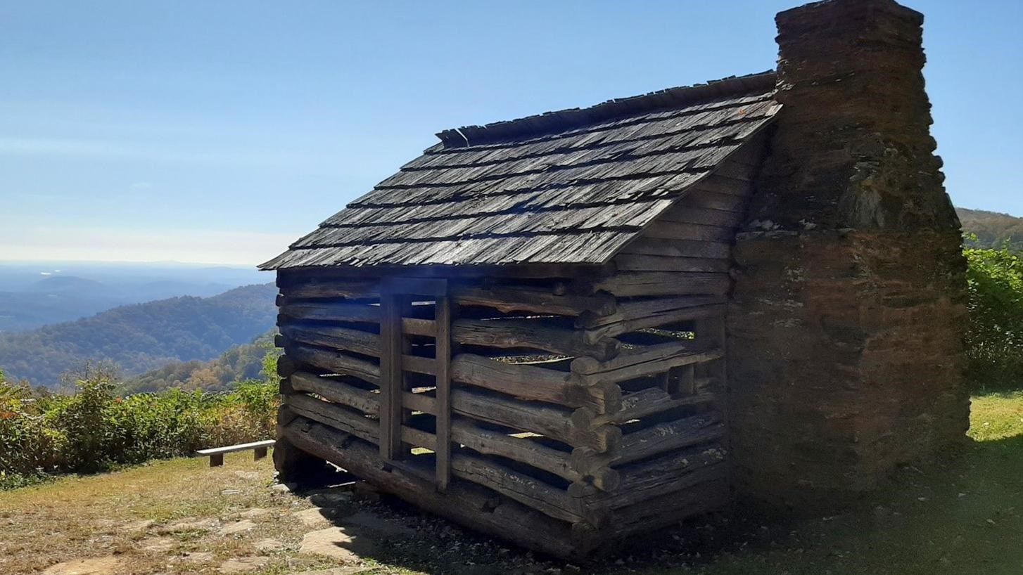A 12 by 12 log cabin with gaps between the boards and a stone chimney sits atop a hill.