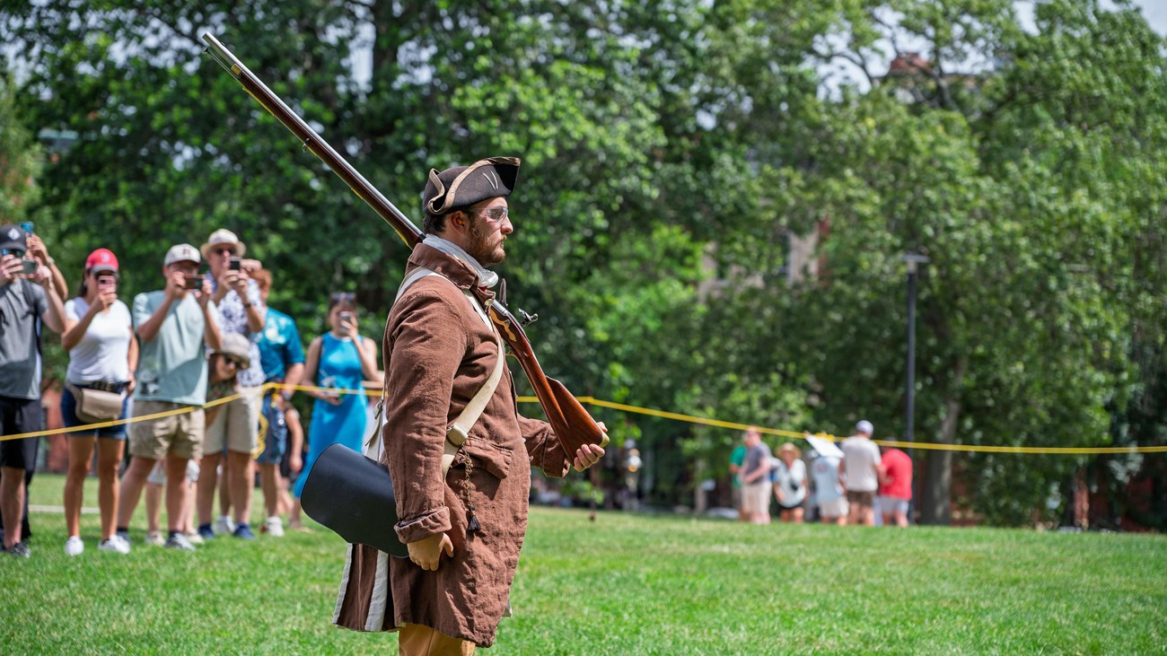 A ranger dressed in period clothing has a musket resting on his shoulder in front of an audience
