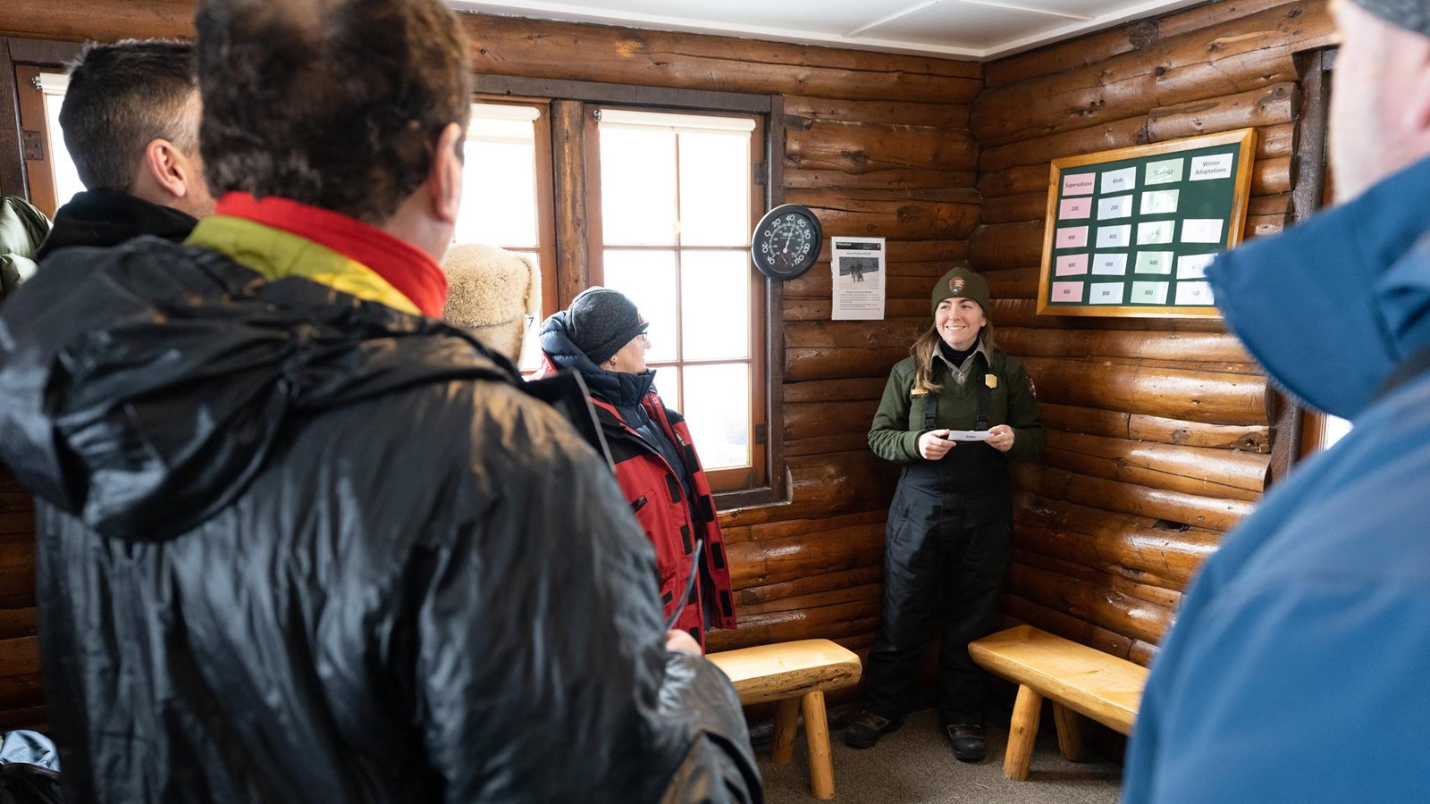 A ranger speaks to visitors in snowmobile suits inside a log building