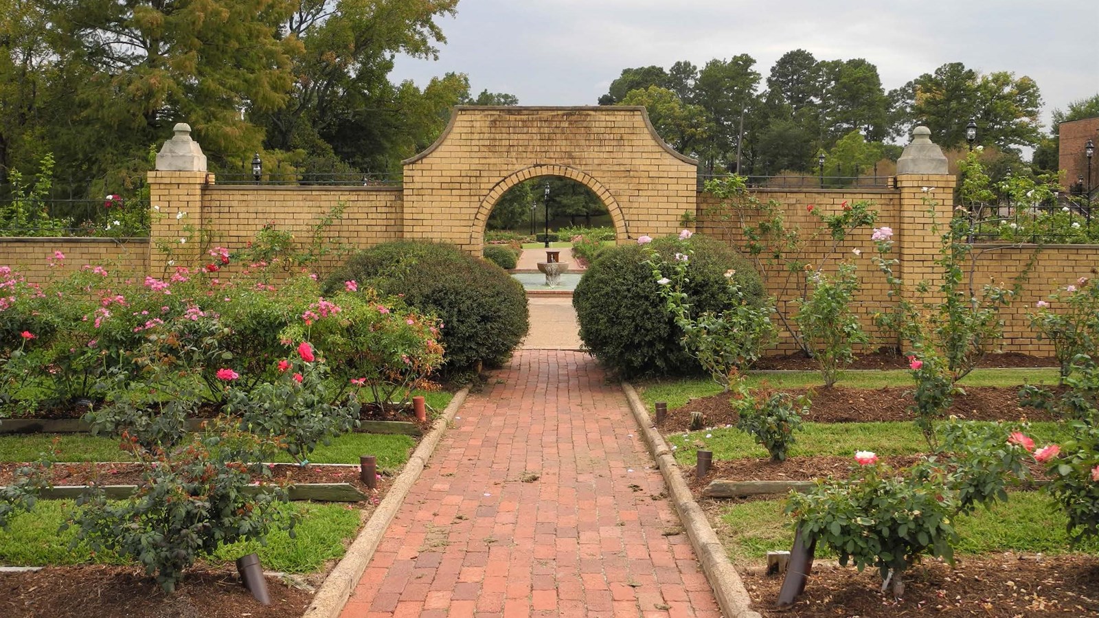 path leading to arched opening in a brick wall, arranged rose beds on either side of path