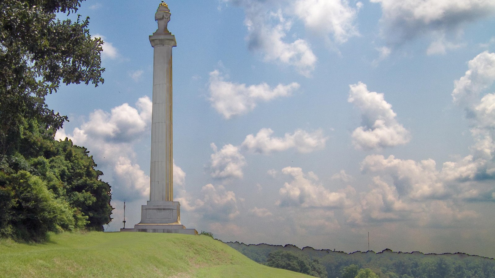 A green grassy ridge with a tall column monument and trees on top with white clouds and blue sky.