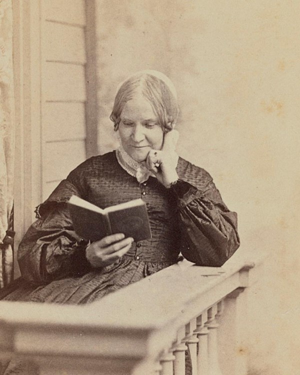An older woman sits with her hand on her chin, leaning on a balcony while reading a novel 