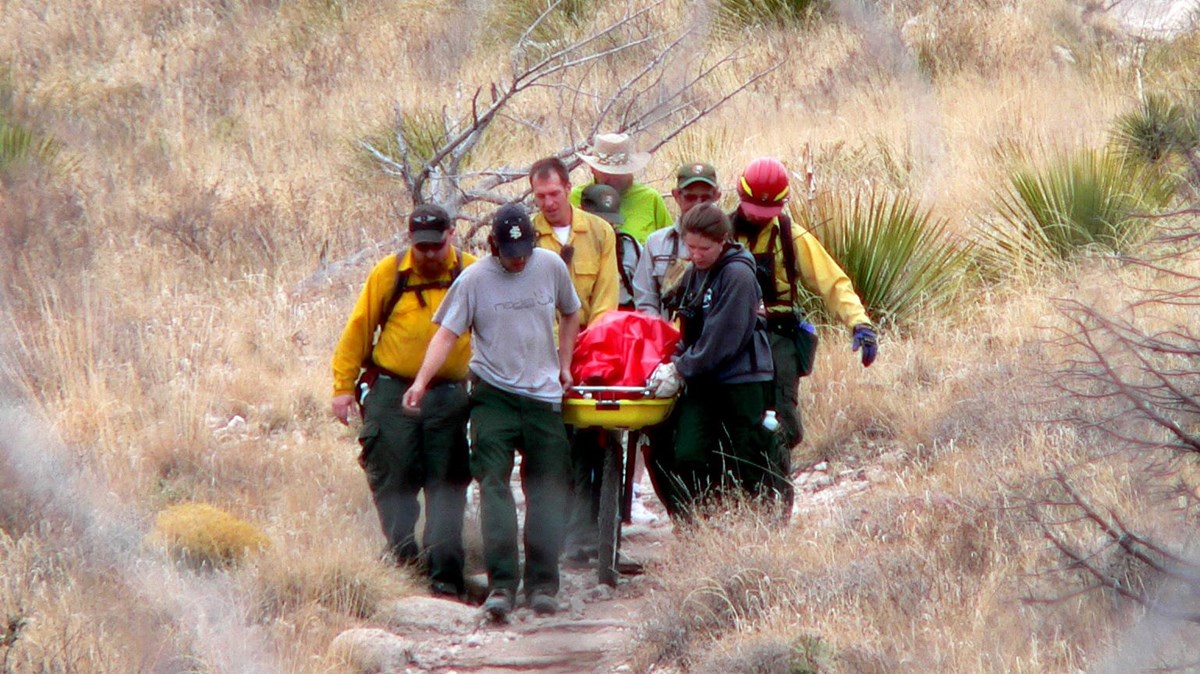 A group of uniformed staff carry a wheeled litter down a rocky trail in a desert mountain terrain