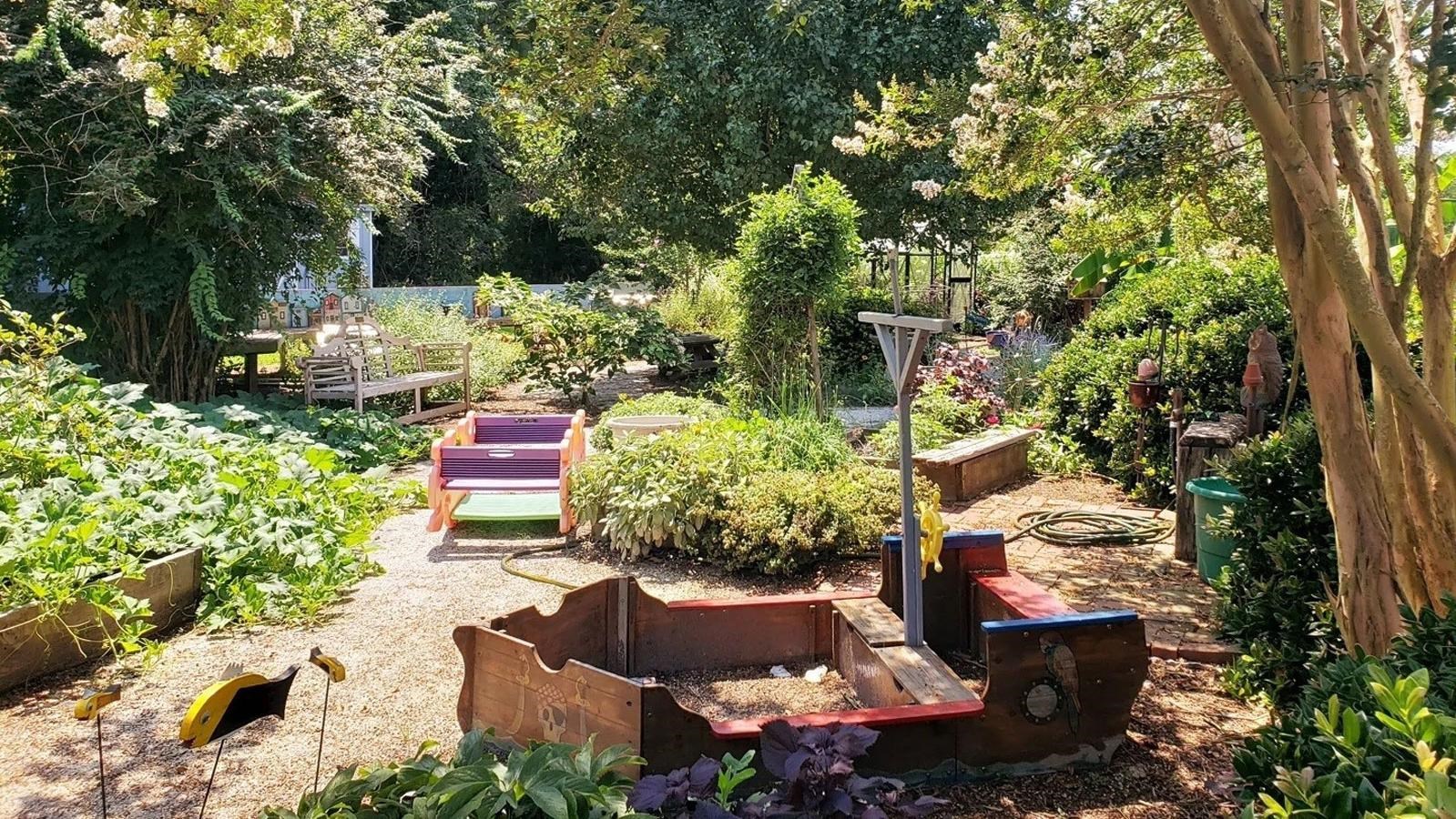 A sunlit garden with a sandbox shaped like a boat, surrounded by lush greenery and trees.