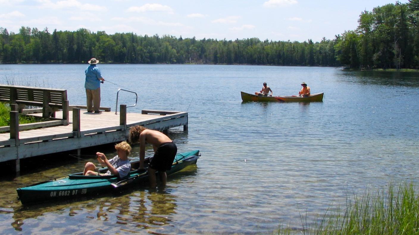 A woman fishes from the dock, canoeists paddle by, kayakers unload along the shore