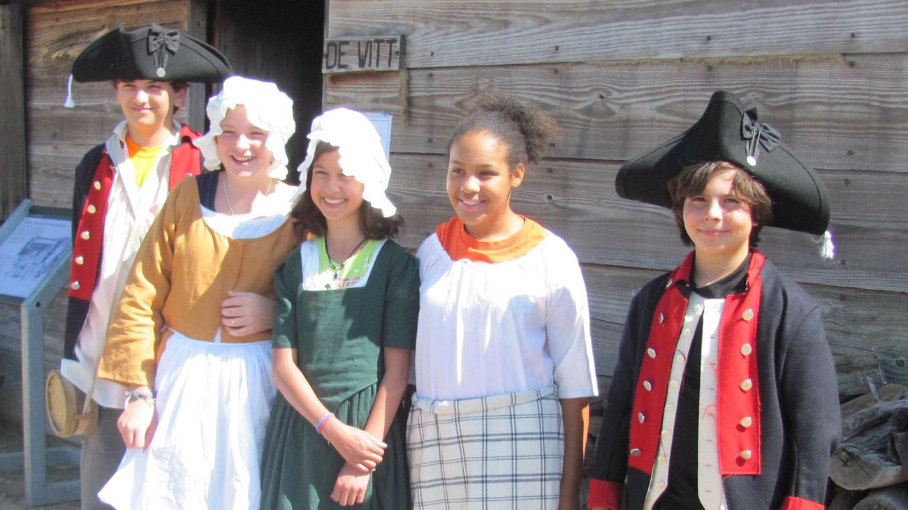 Five children stand in a row smiling. They all have different 18th Century accessories on.