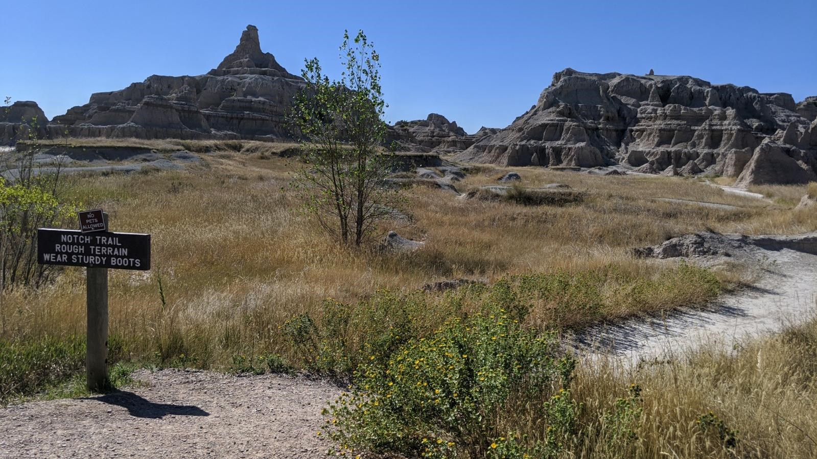 A sign marked Notch Trail alongside a trail proceeding amid badlands buttes under blue sky.