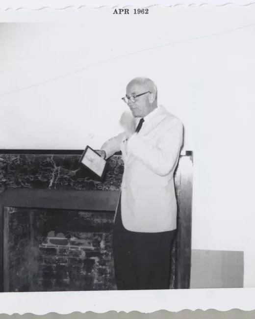 Black and white image of a man speaking while leaving on a fireplace mantel. 