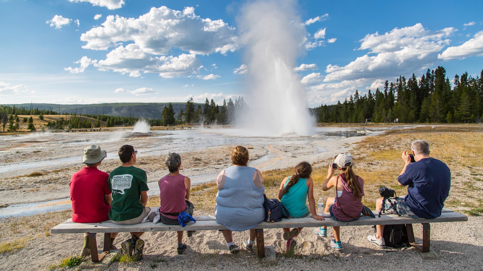 People sit on a bench and watch a geyser shoot water about 75 feet into the air at a slight angle.