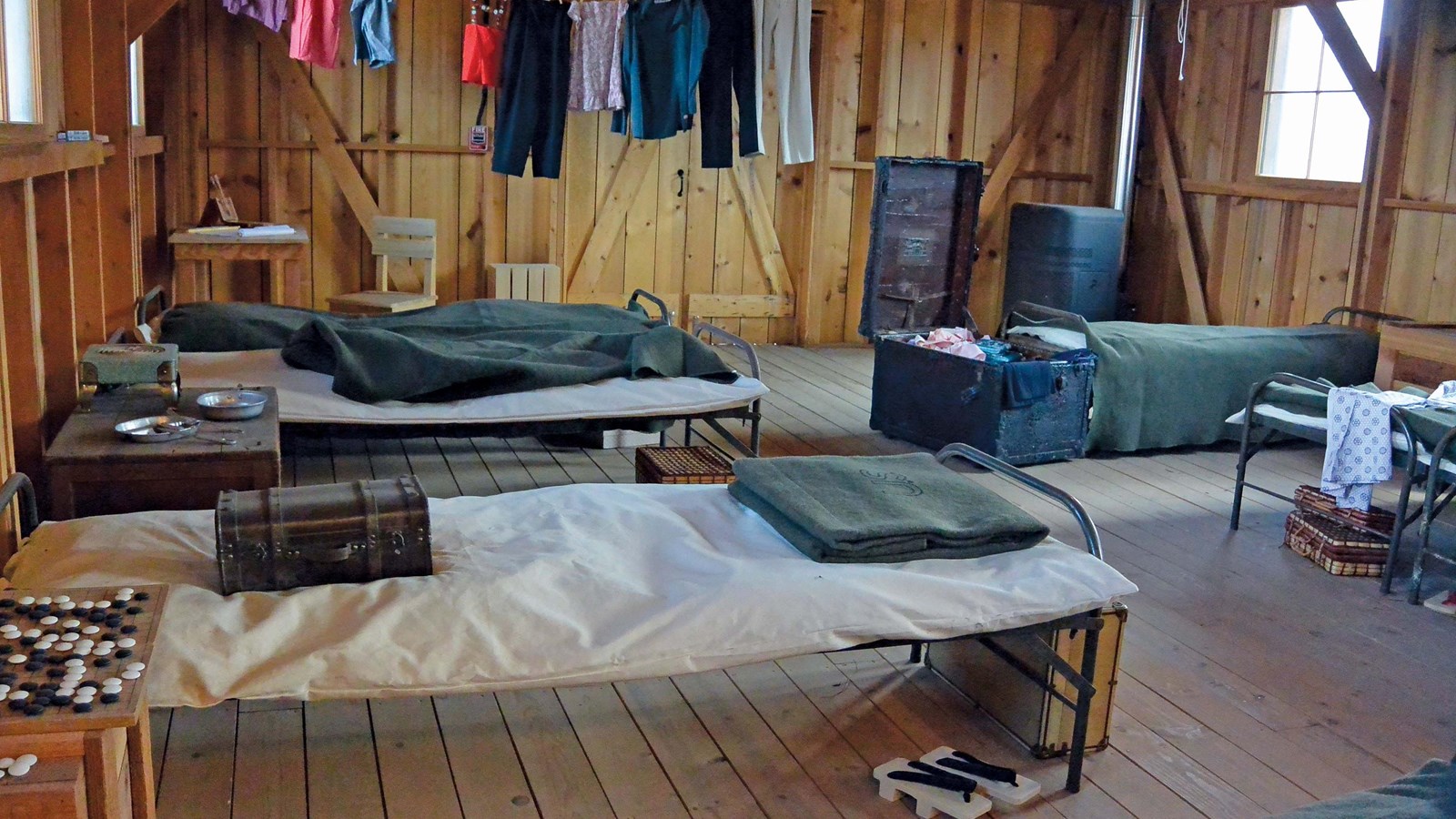 Army cots line the walls with army blankets. Clothes is hung along the back wall.