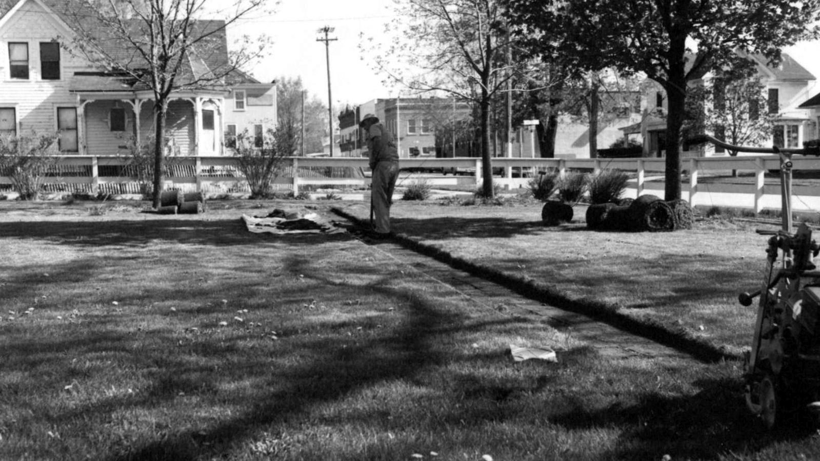 A man is in the process of digging a trench in a vacant grassy lot in preparation of an archeologica