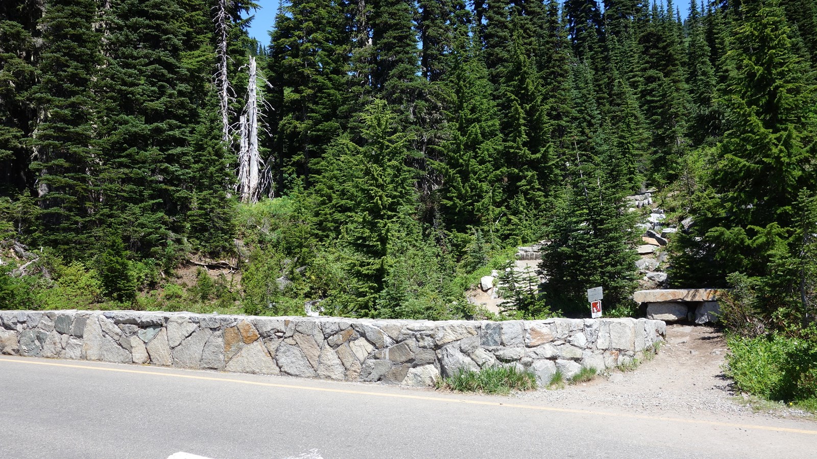 A trailhead starts next to a low rock wall along a paved road. 