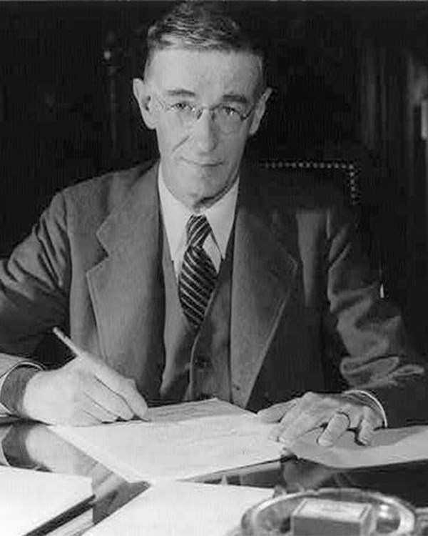 A black and white photo of a slender man in a suit, seated at a desk with numerous documents.